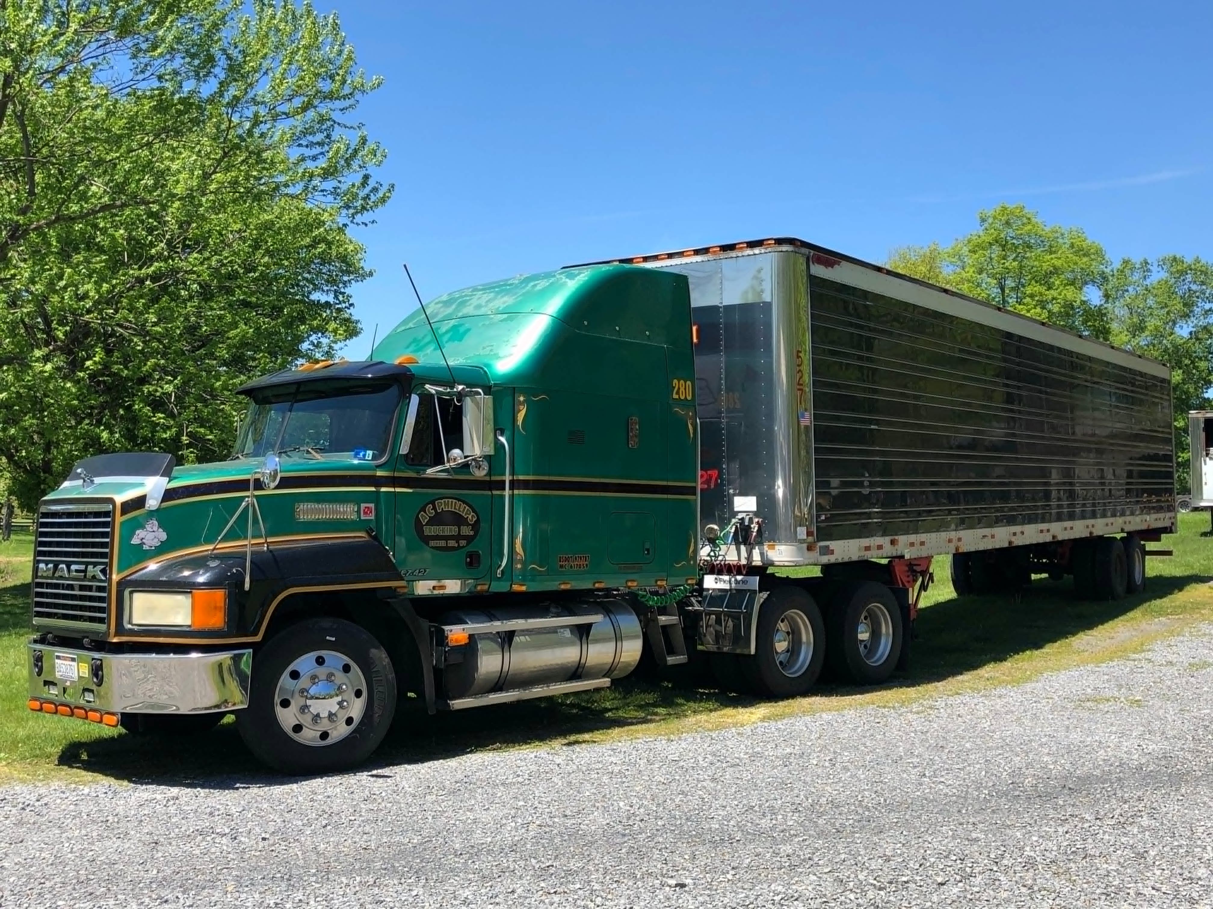 A green and black Mack semi-truck with a chrome trailer parked on a gravel path surrounded by lush green trees and grass under a clear blue sky.